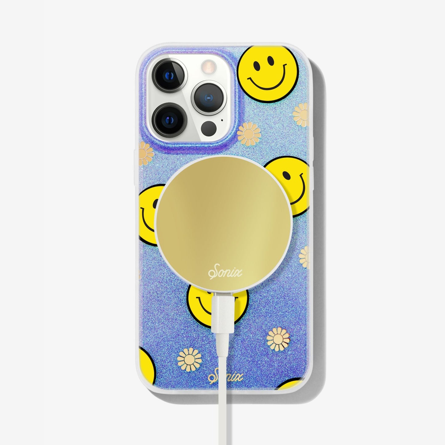 The clear purple case infused with iridescent glitter features happy faces and signature gold foil flowers shown on an iphone 12 with a gold maglink charger on the back