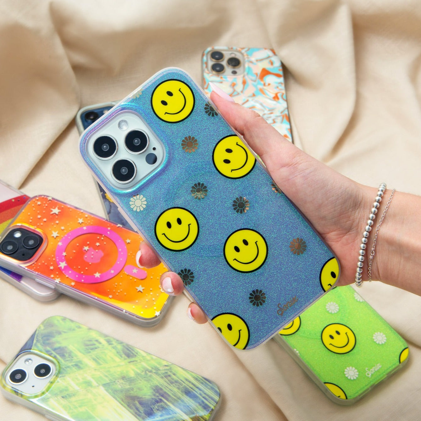 The clear purple case infused with iridescent glitter features happy faces and signature gold foil flowers shown on an iphone being held by a hand and other phone cases in the background