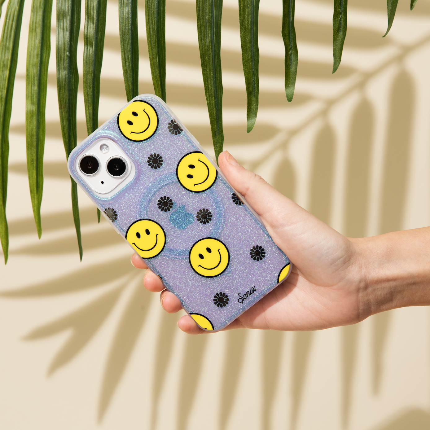 The clear purple case infused with iridescent glitter features happy faces and signature gold foil flowers shown on an iphone with a hand holding it infront of a palm tree
